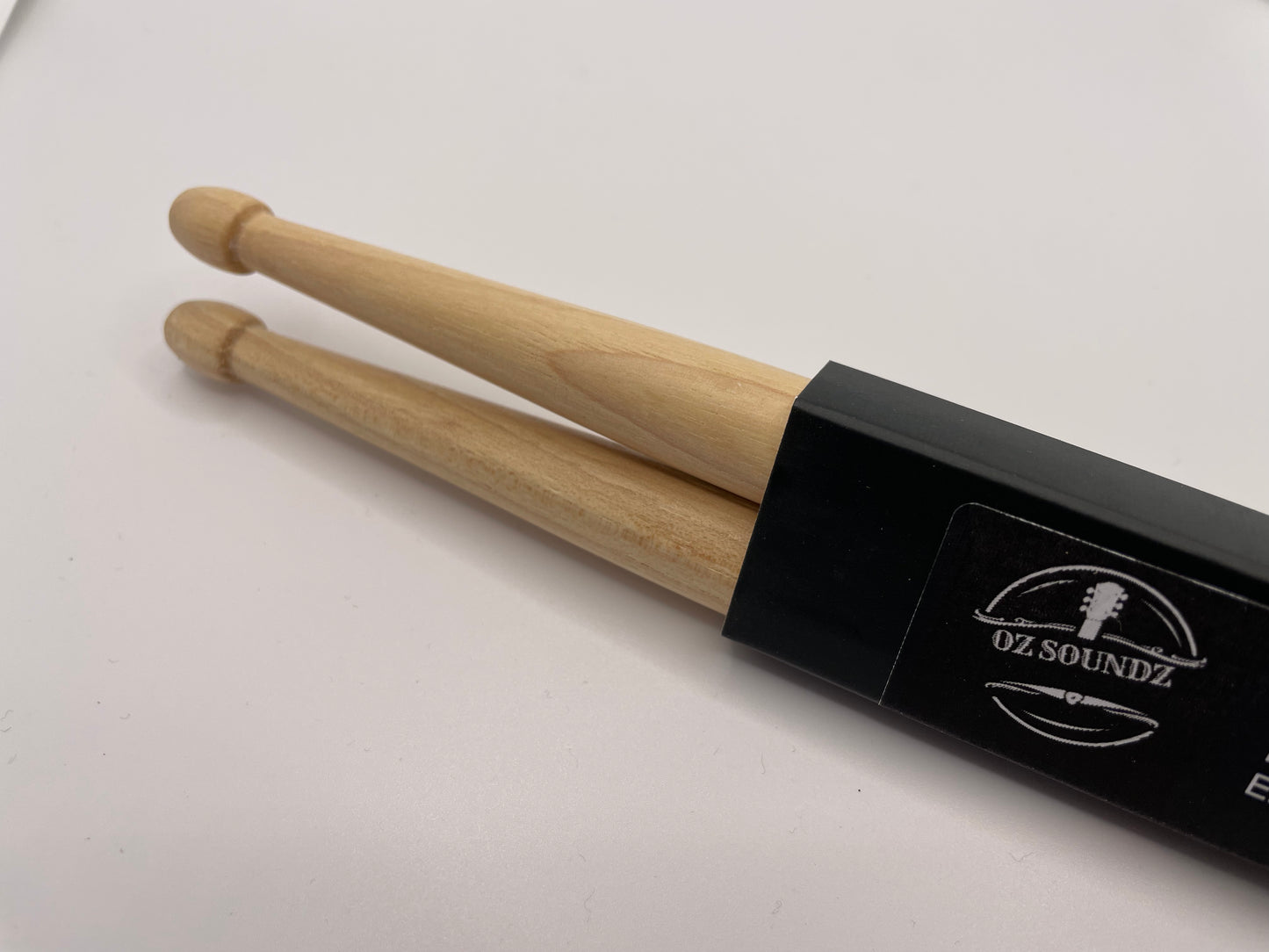 A pair of North American Hickory wood drumsticks with a Acorn Tips in black Oz Soundz packaging, placed on a white surface.